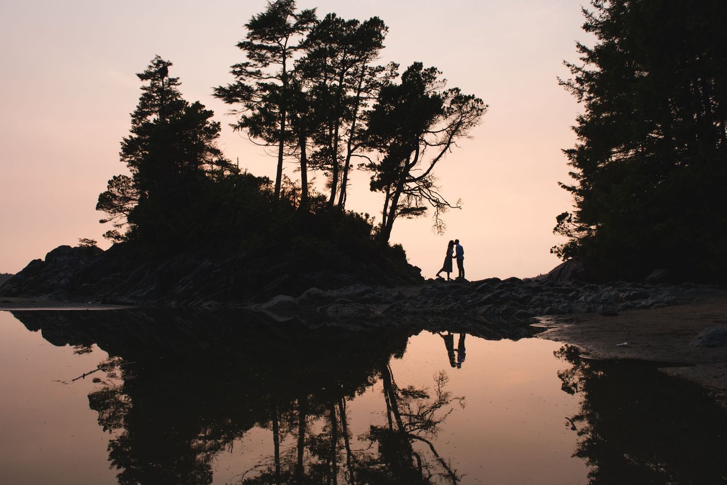 Scenic sunset view over a rocky lake shore with silhouetted pine trees against a pink and orange sky.