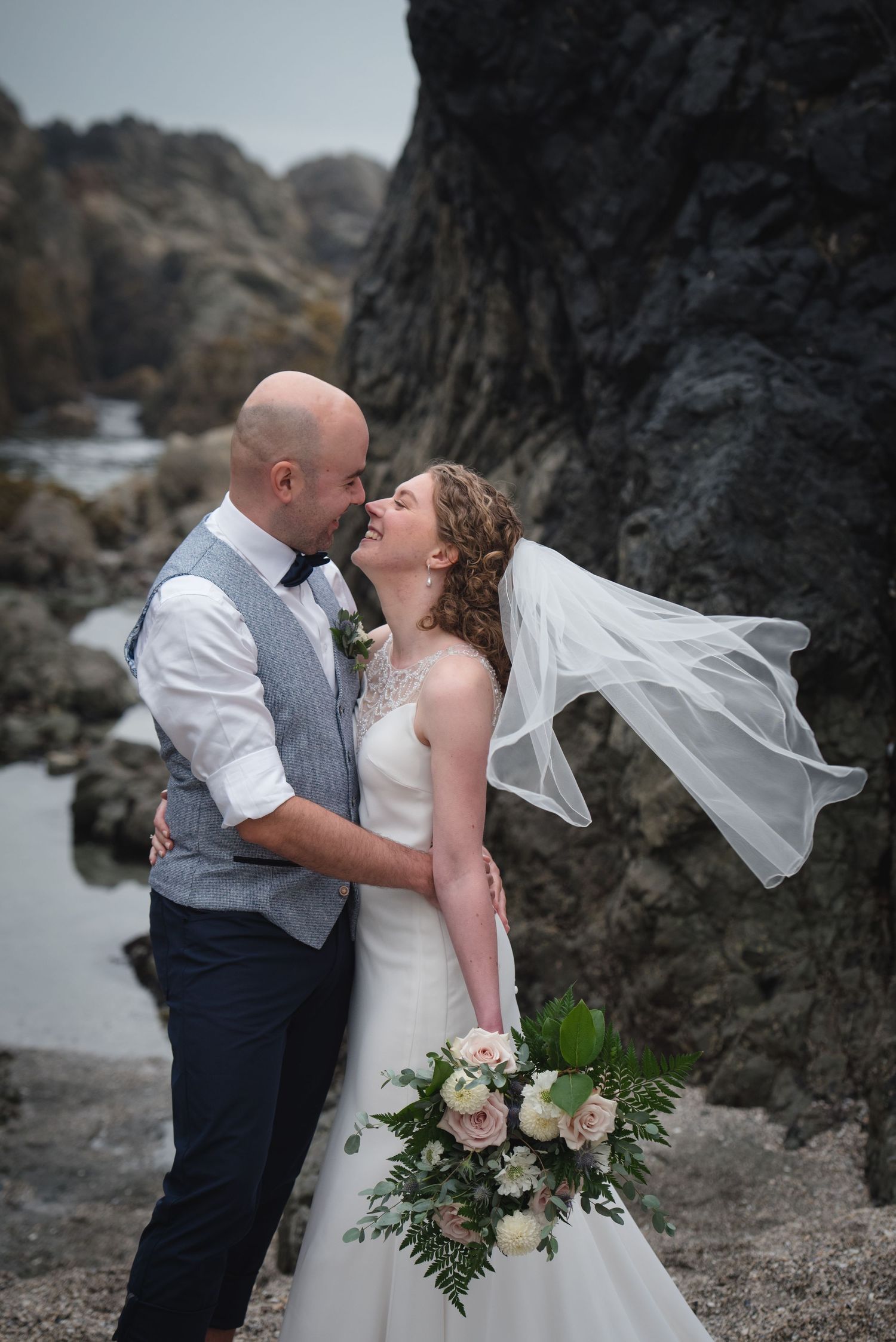 A couple shares an intimate moment on a dramatic rocky coastline while holding a pink and white floral bouquet.