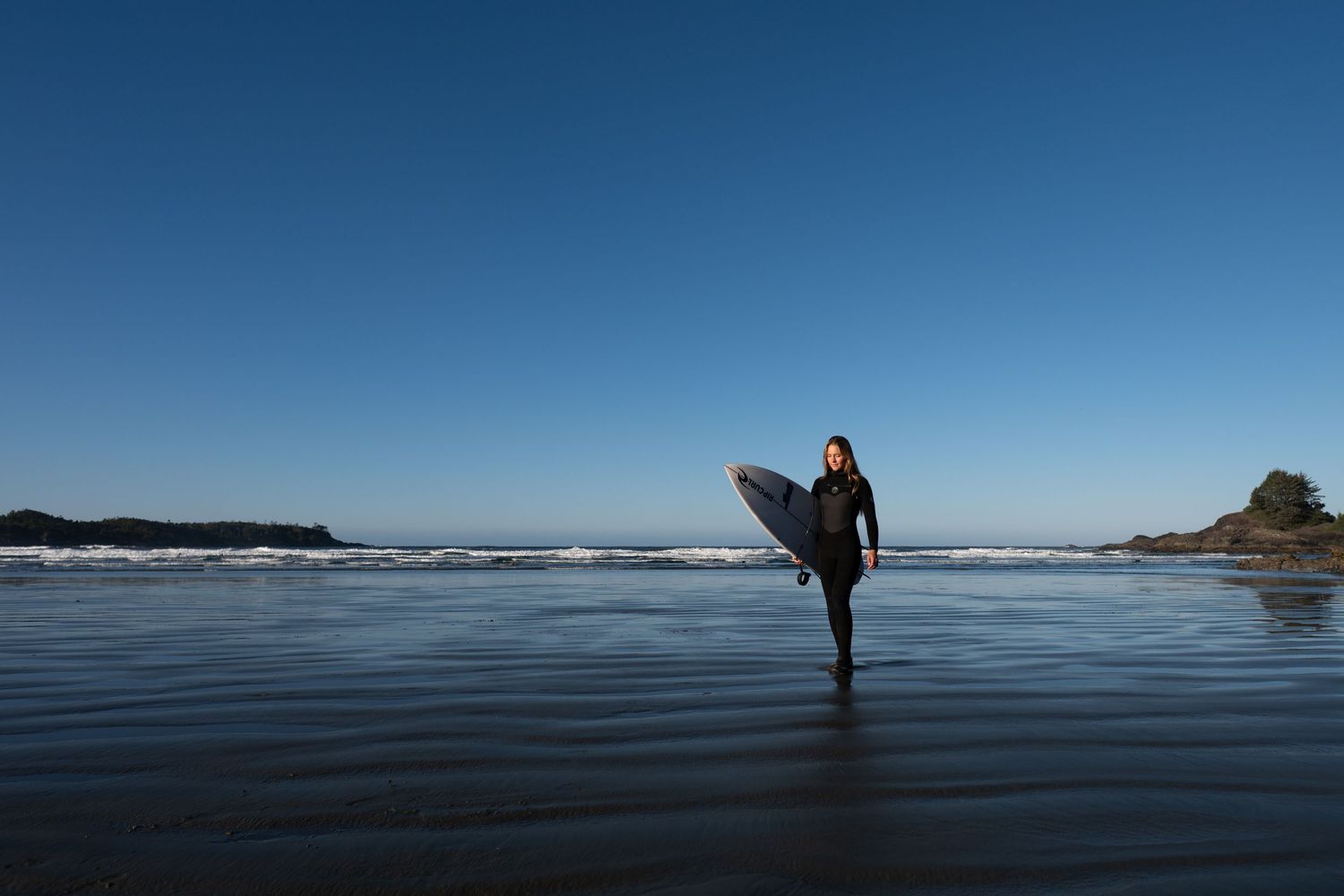 Silhouette of surfer walking along misty Pacific Northwest coastline at sunset with mountains in background.