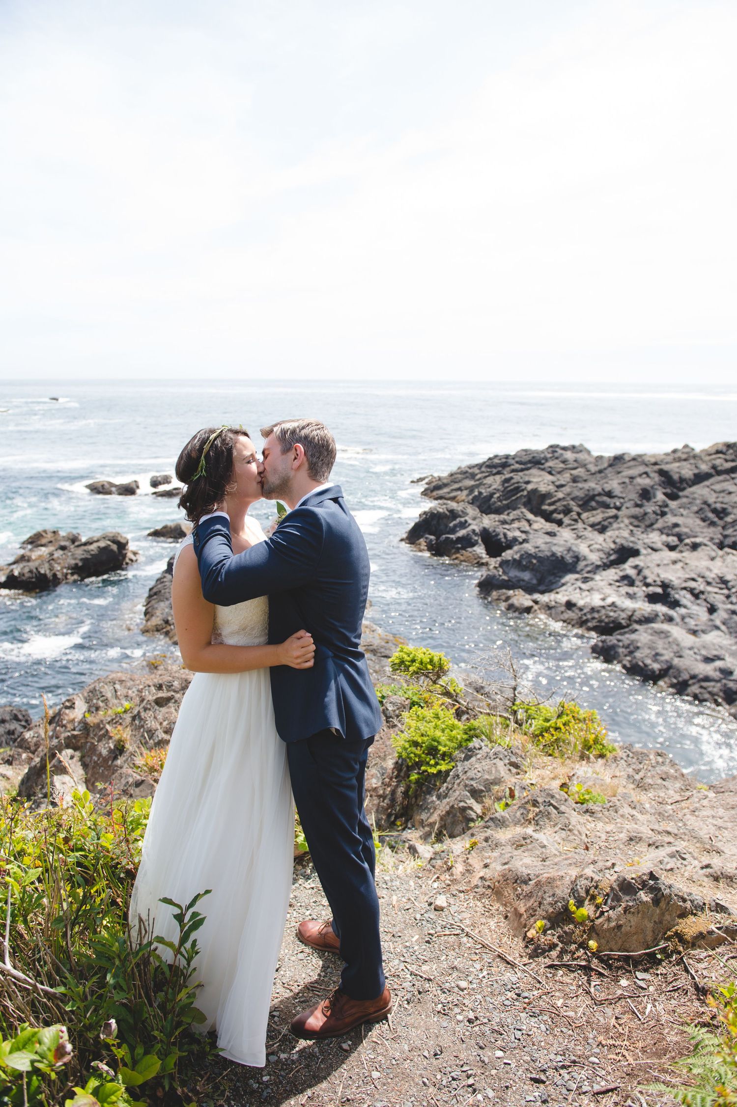 Romantic coastal wedding photos with couple embracing on rocky cliff overlooking the ocean.