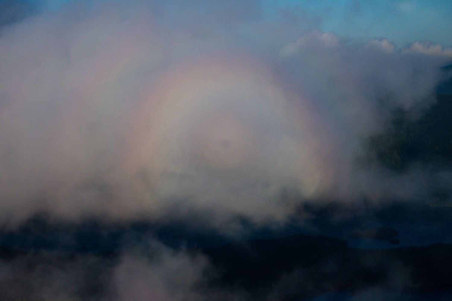 Beautiful full circular fogbow rainbow glowing in misty clouds above dark mountain landscape at dusk.