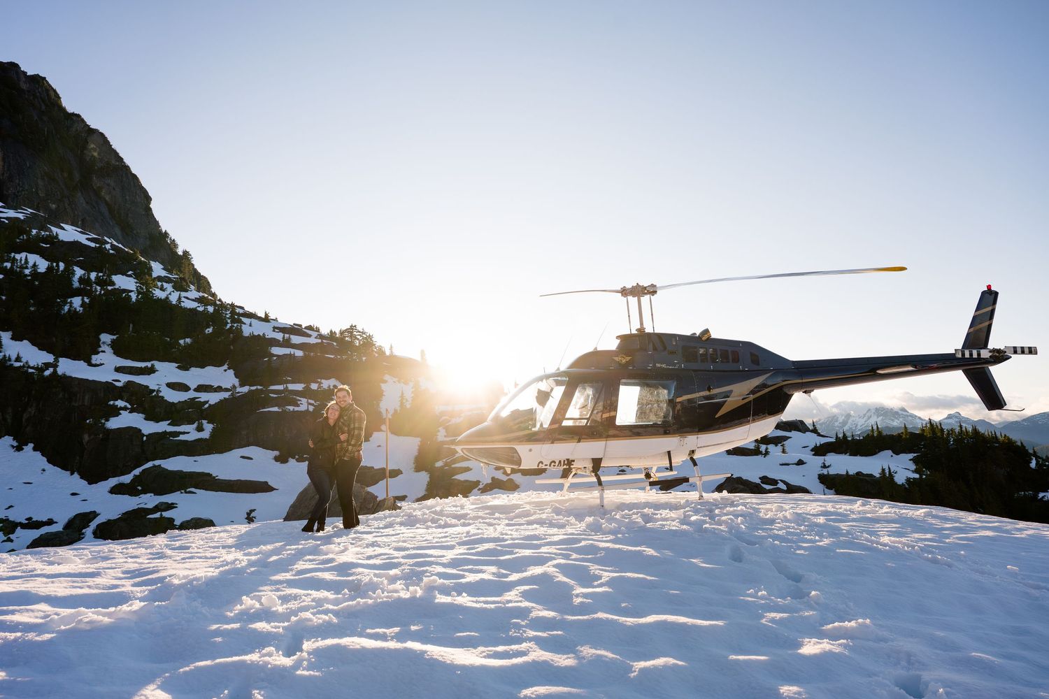 Helicopter landed on snowy mountain in Tofino, backlit by bright sunlight in a winter alpine setting.