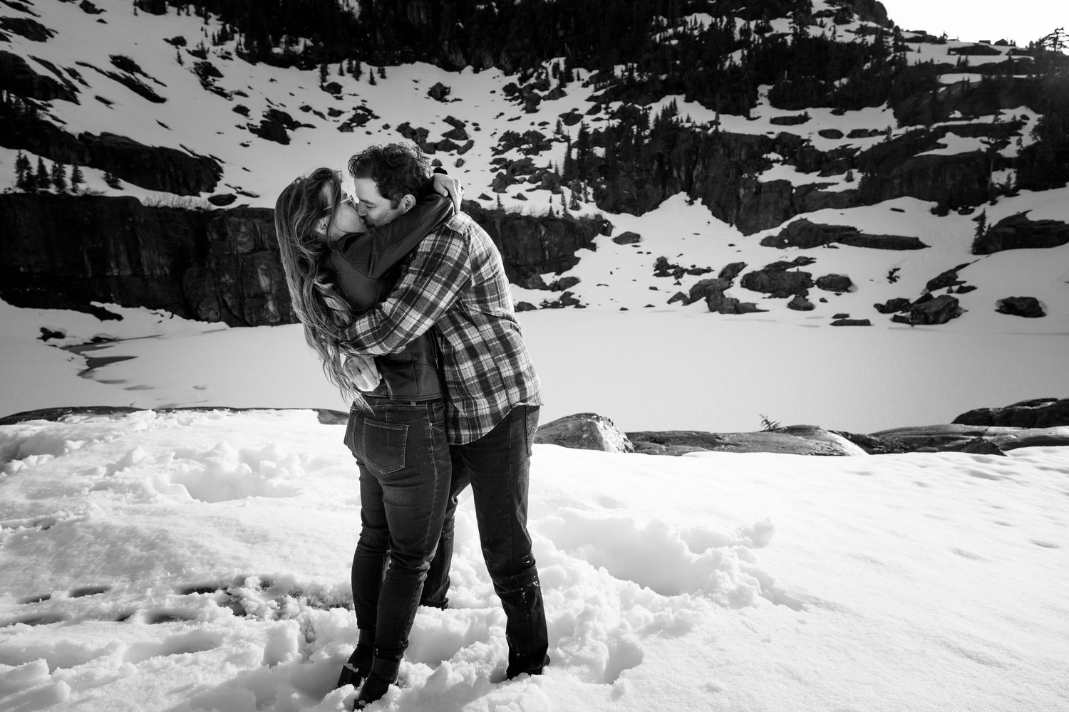 Couple embracing and kissing in snowy mountain landscape, black and white winter engagement photo.