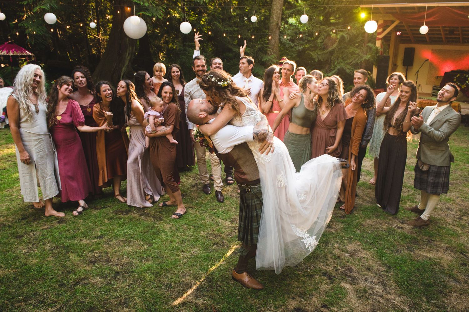 Outdoor wedding reception with string lights and paper lanterns illuminating guests dancing and celebrating at dusk.