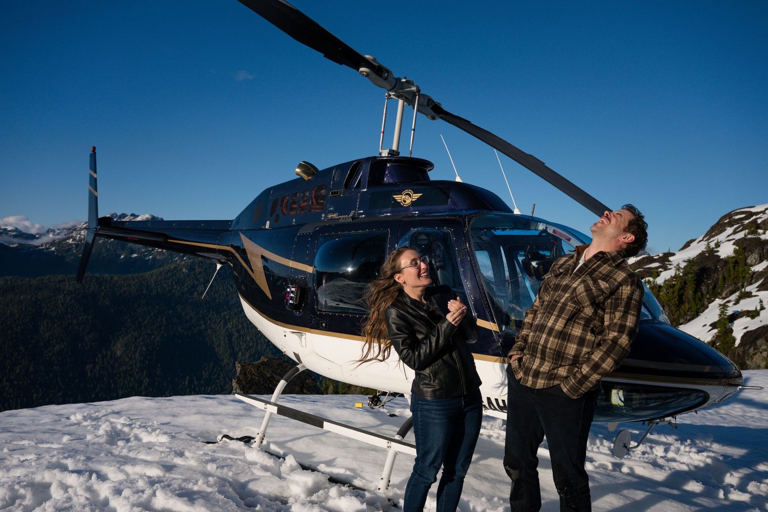 Couple laughing together beside an Atleo Air helicopter on a snowy mountain peak under a clear blue sky.