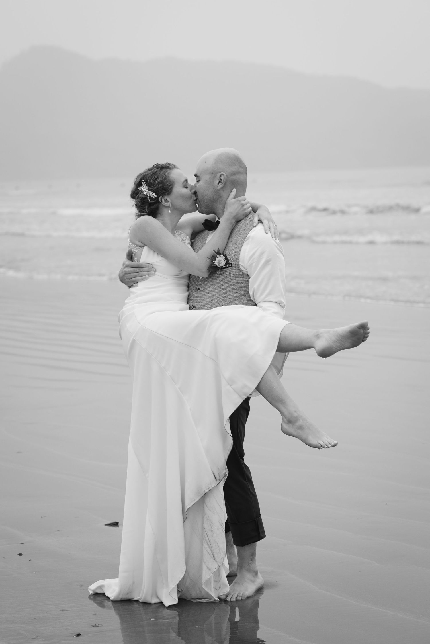 Black and white photo of newlyweds embracing playfully at the water's edge on a beach.