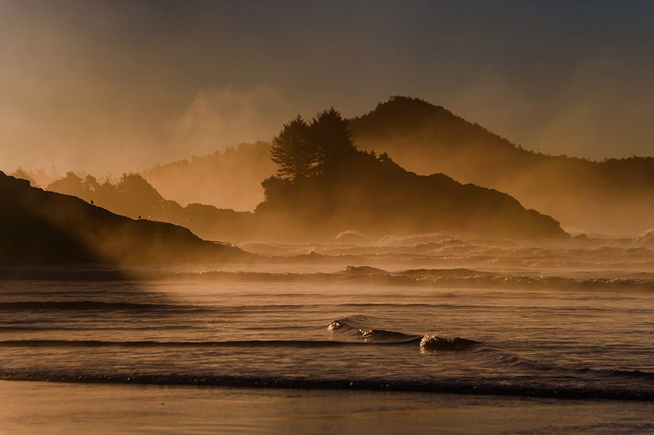 Misty coastal landscape at sunset with mountains silhouetted against golden sky.