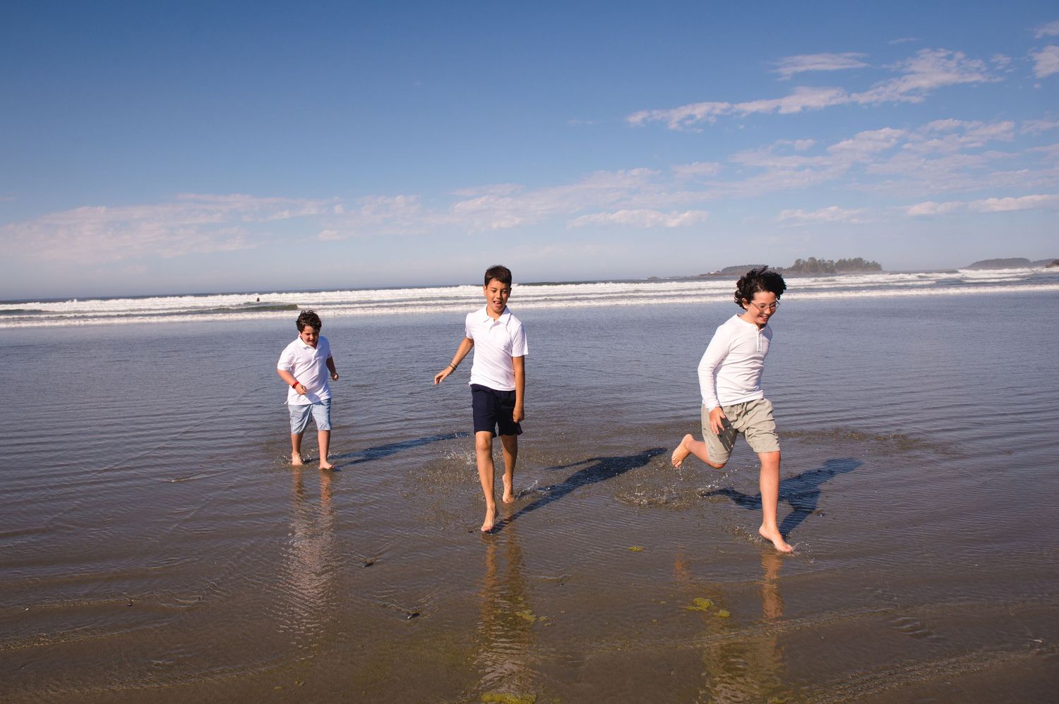 Three people running and splashing in the shallow ocean water on a sunny beach day.