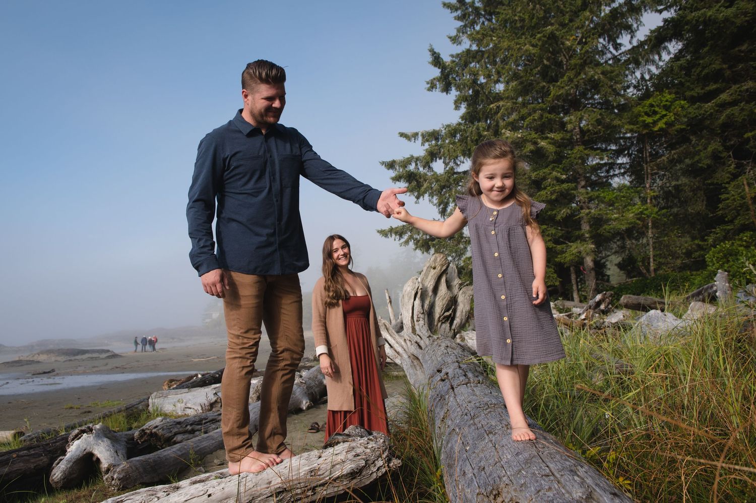 Casual outdoor family photography session on a beach with driftwood and trees.
