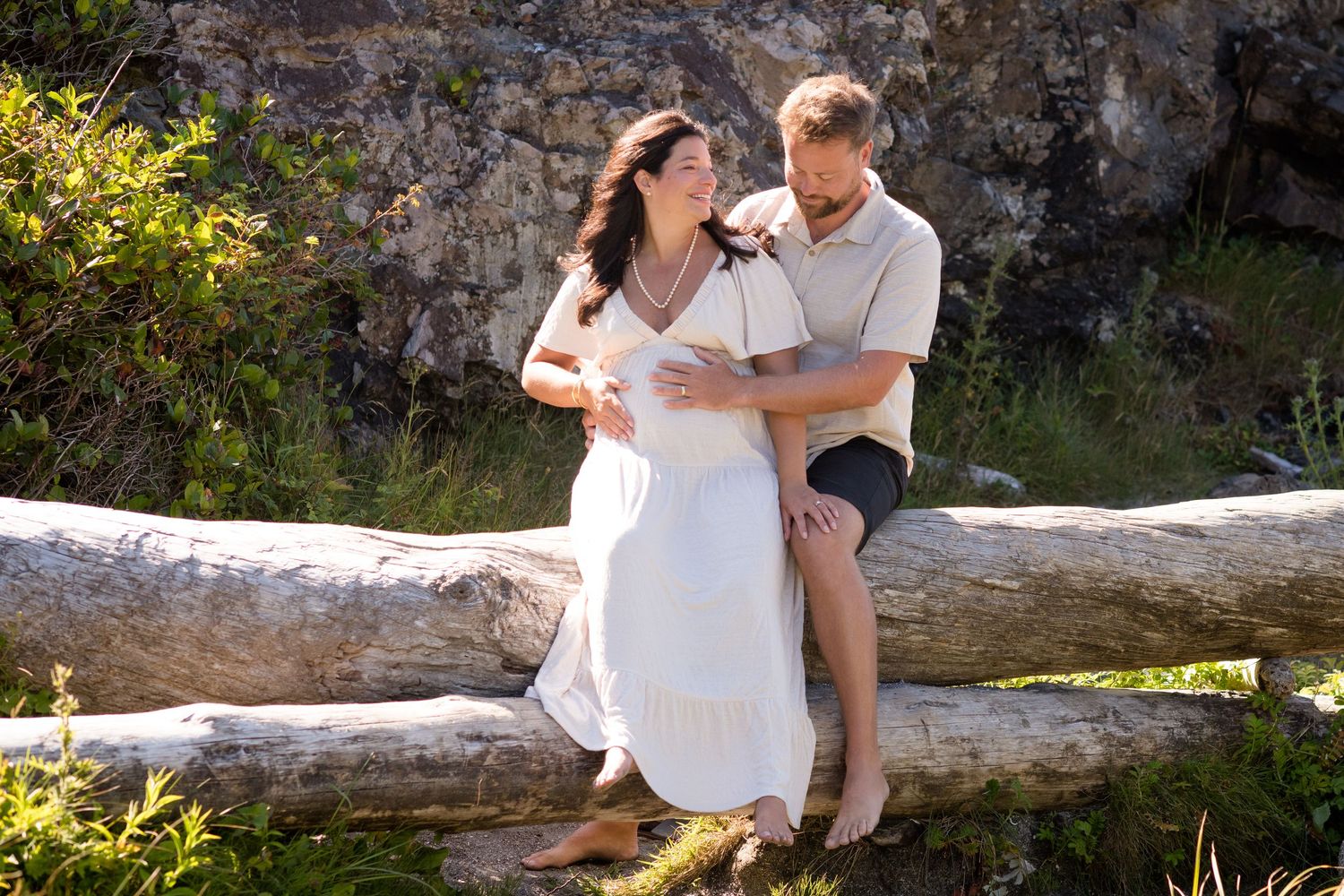Two people embrace while sitting on a large driftwood log against a rocky coastal backdrop.