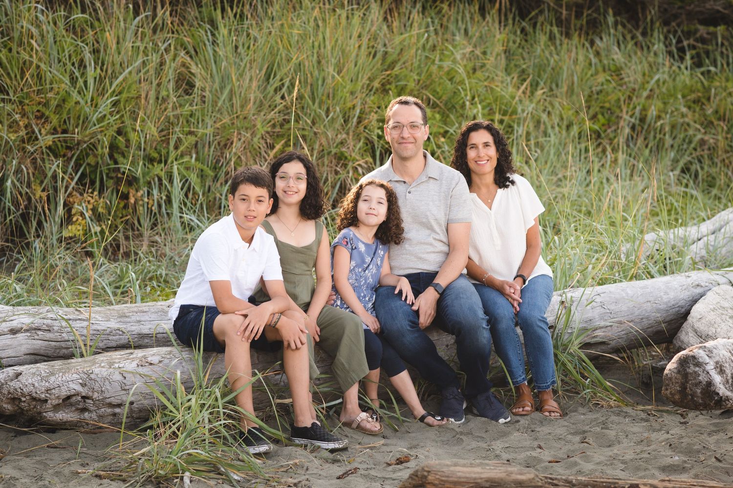 Family relaxing on beach logs with tall grass and natural scenery in the background.