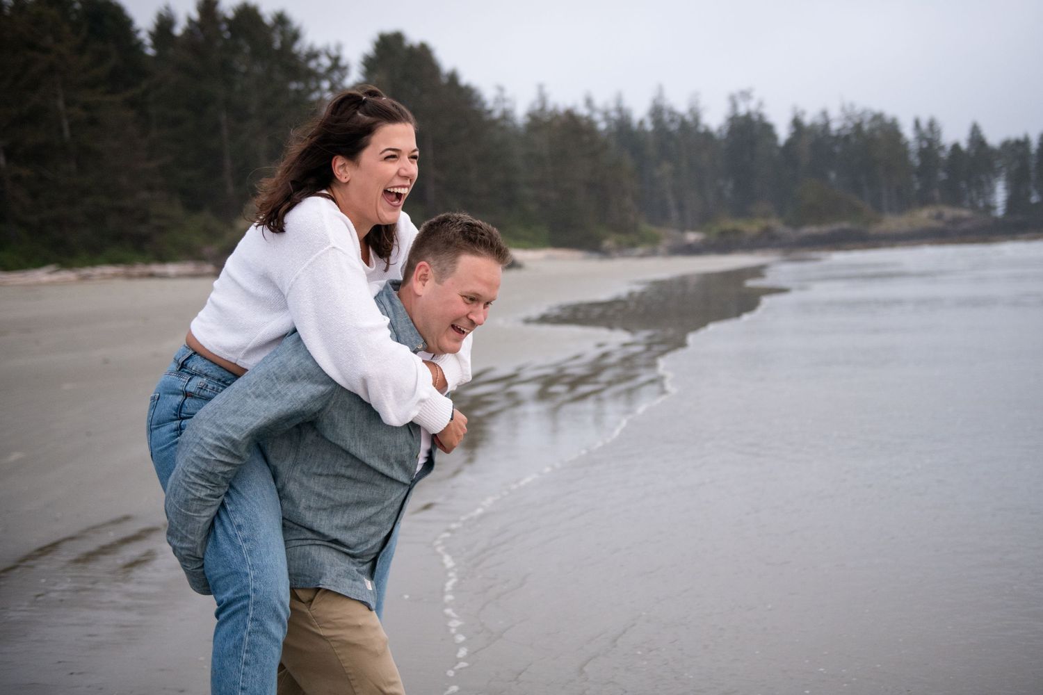 A playful couple frolics on a misty Pacific Northwest beach with evergreen trees in the background.