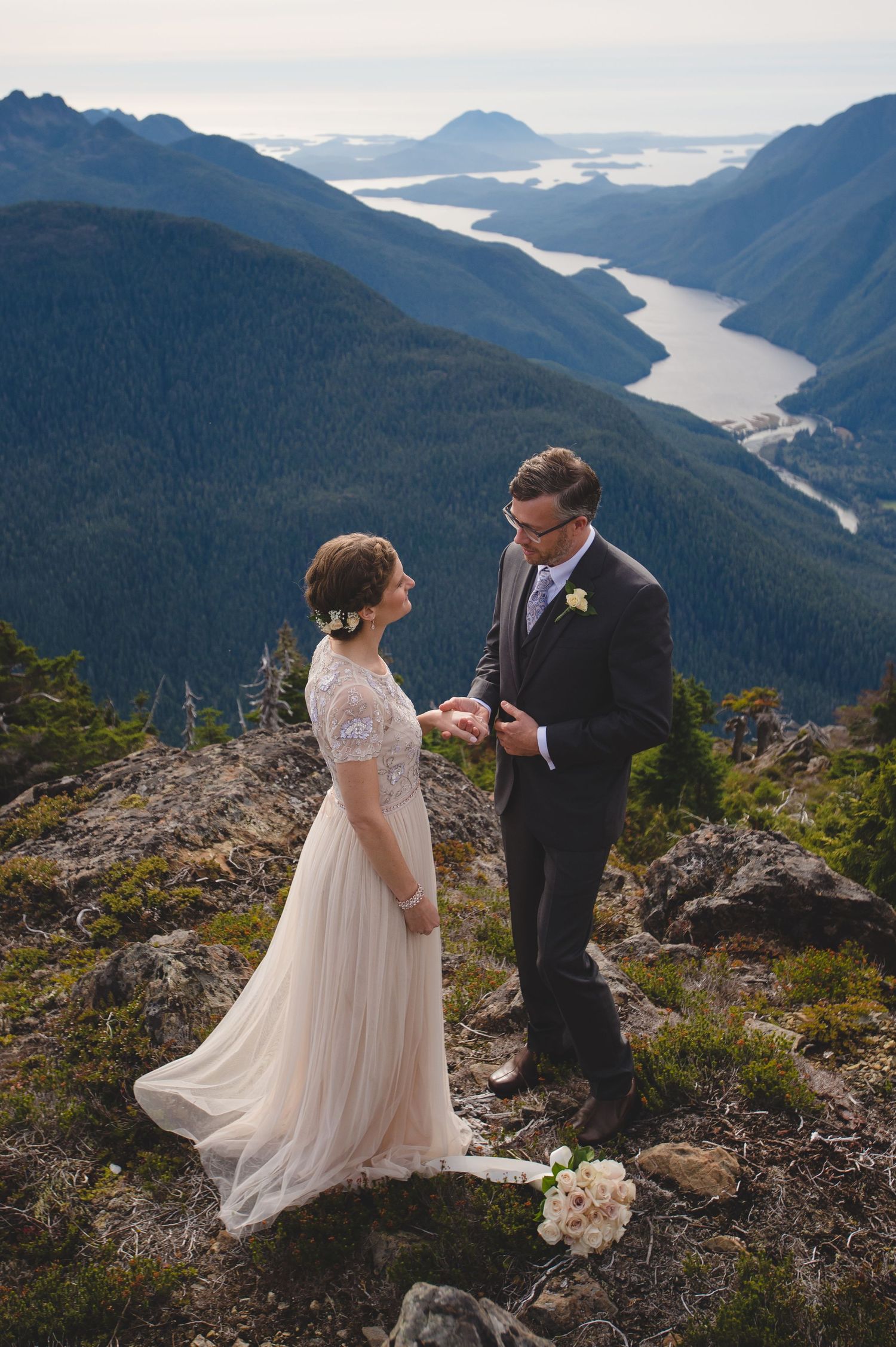 A couple in wedding attire stands on a rocky mountain peak overlooking a dramatic fjord landscape.