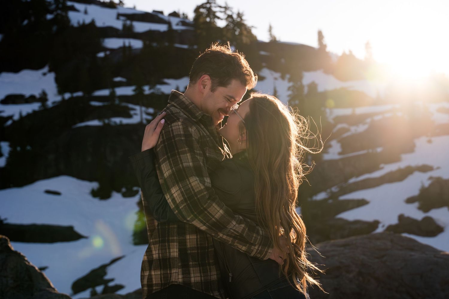 Couple sharing a romantic kiss at golden hour on a snowy mountain with sun flare and snow-covered rocks.