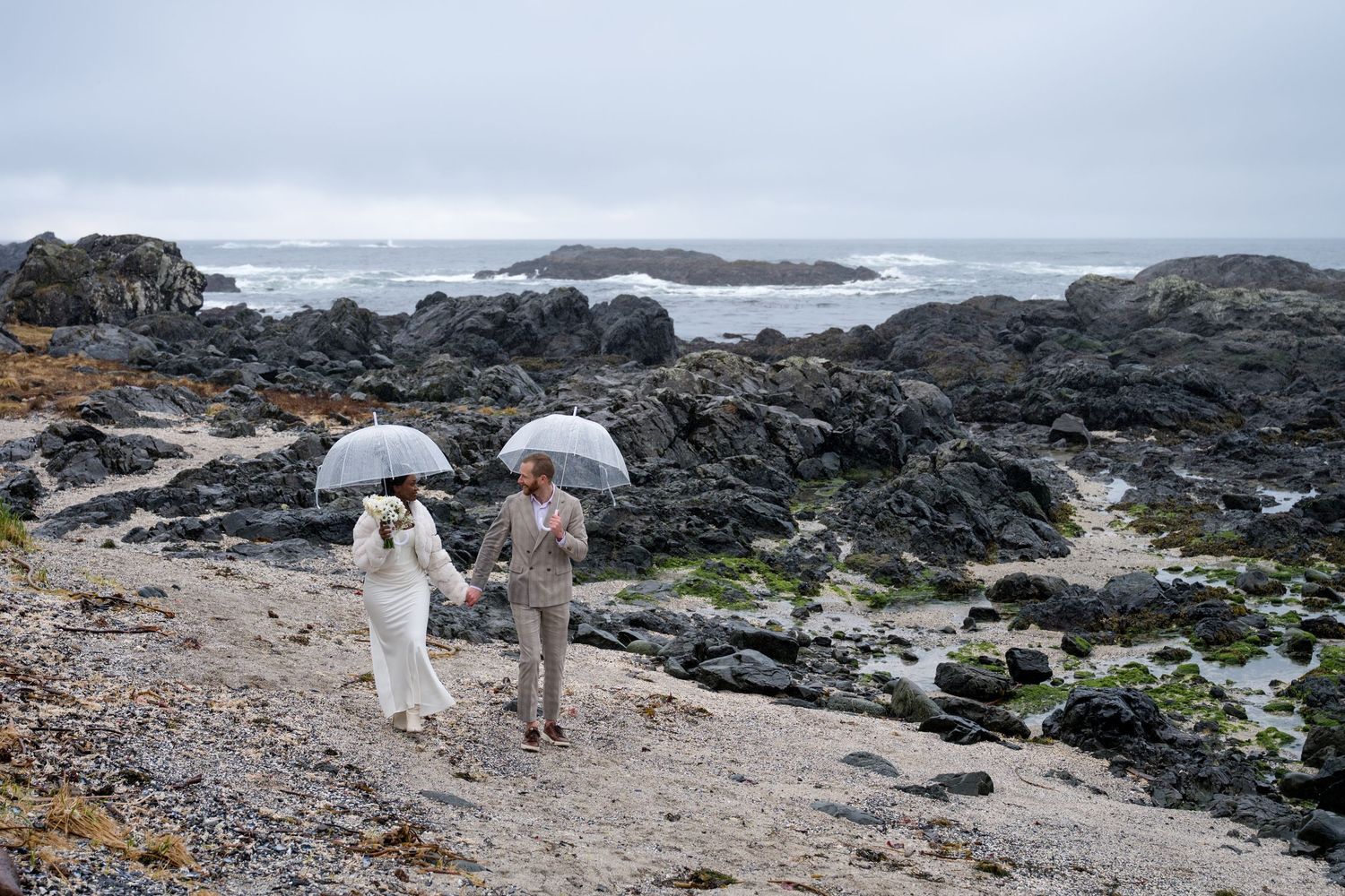 Wedding couple walking along a rocky beach shoreline with dramatic waves and overcast skies.