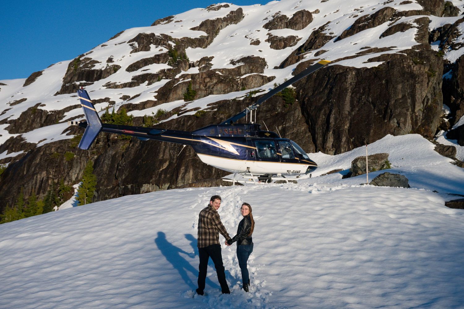 Newly engaged couple holding hands in front of a helicopter on a snowy mountain during a heli proposal.