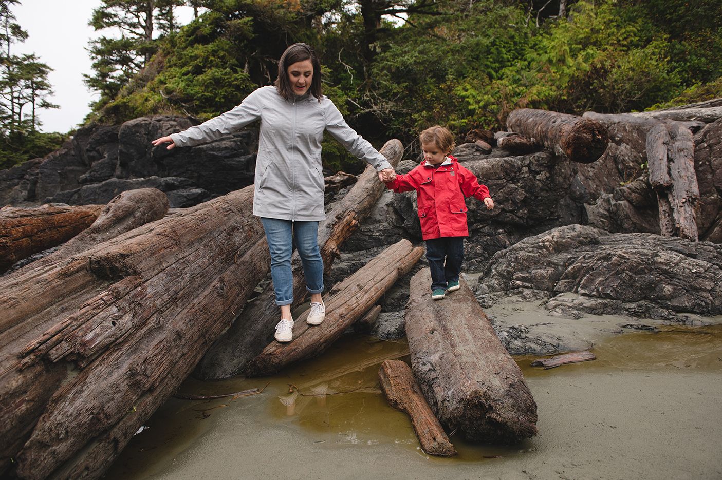 People exploring and climbing on large driftwood logs scattered along a rocky Pacific Northwest beach at low tide.