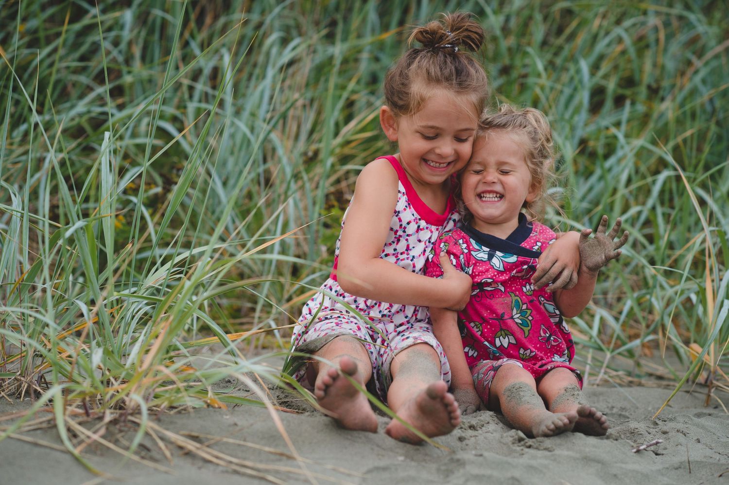 Two young children sitting and laughing together in tall grass on a sandy beach.