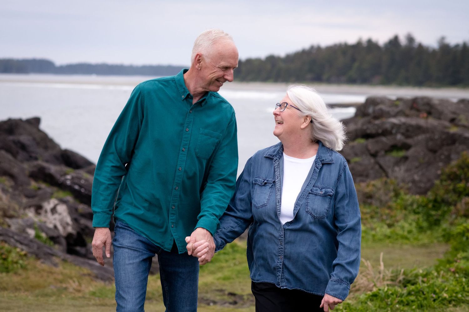 An elderly couple holding hands while walking along a rocky coastal shoreline on a cloudy day.