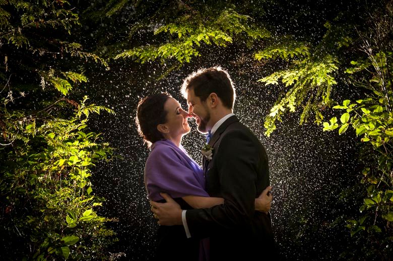 Backlit couple embracing in a forest setting with sunlight streaming through trees.