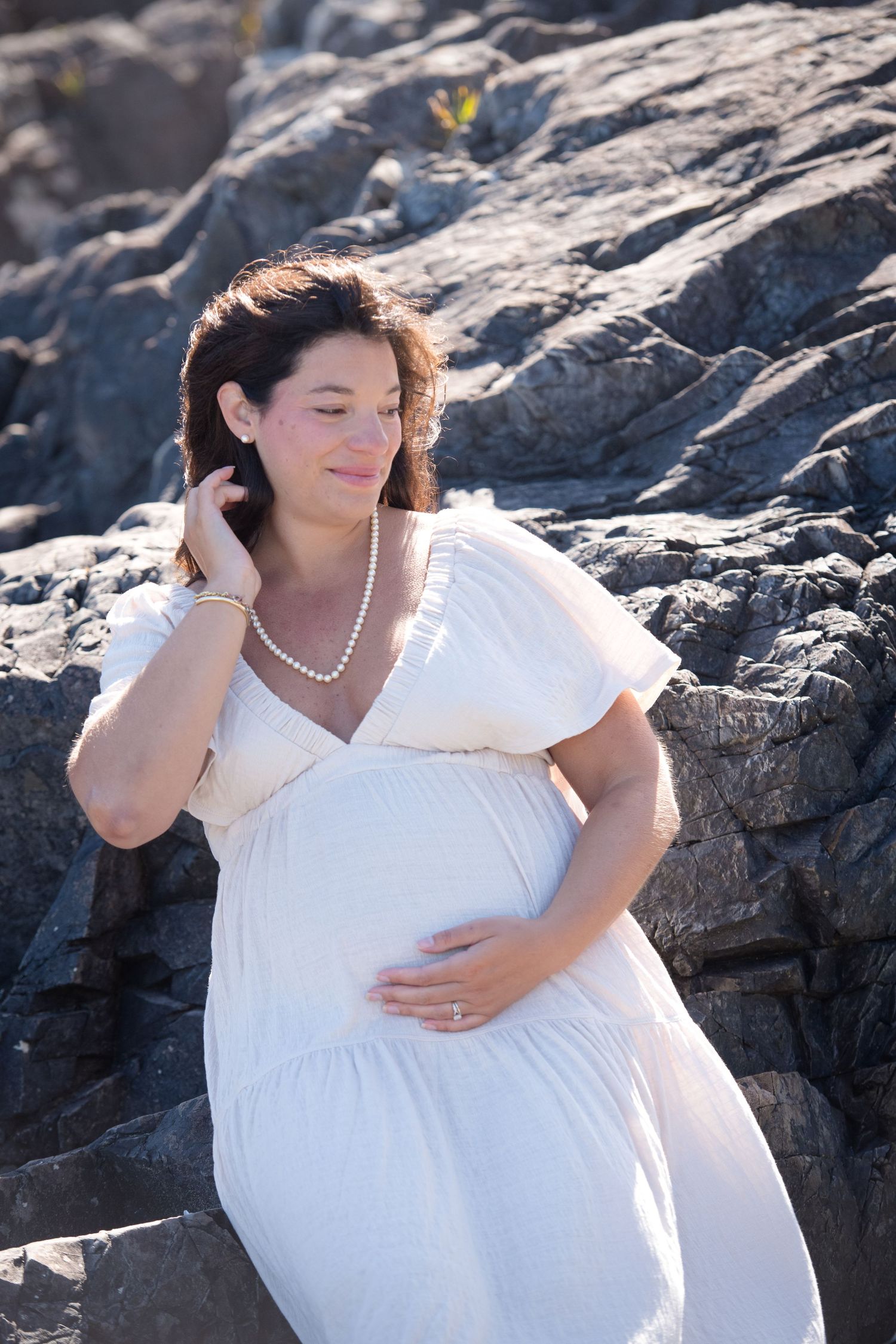 A white dress maternity photoshoot on rocky coastal cliffs during golden hour.