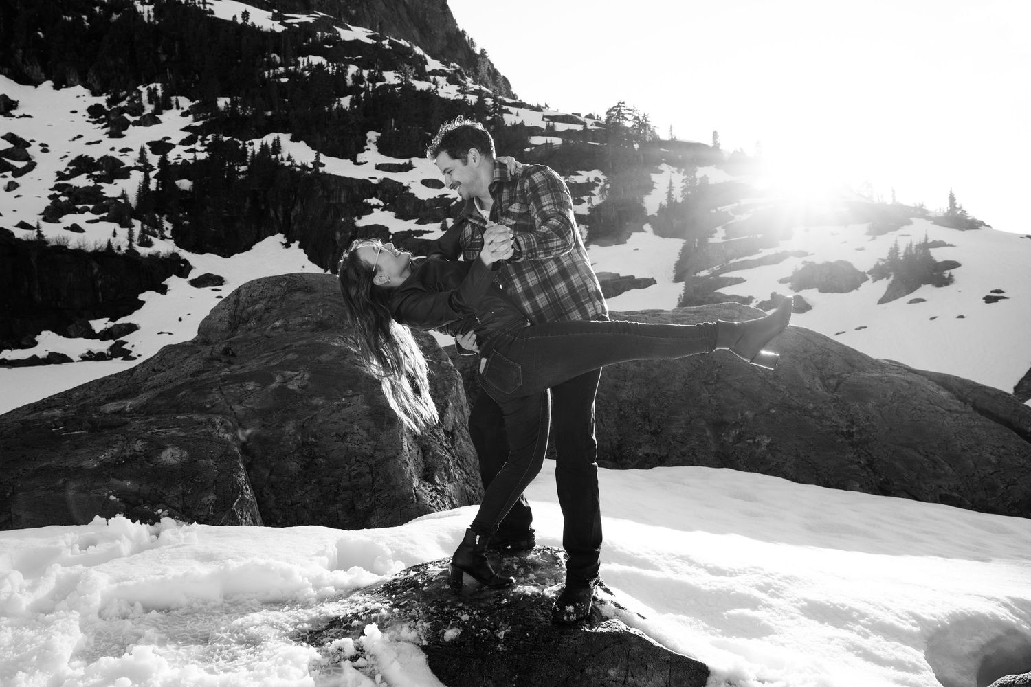 Couple embracing on snowy mountain rocks in black and white photography with bright sunlight backdrop.