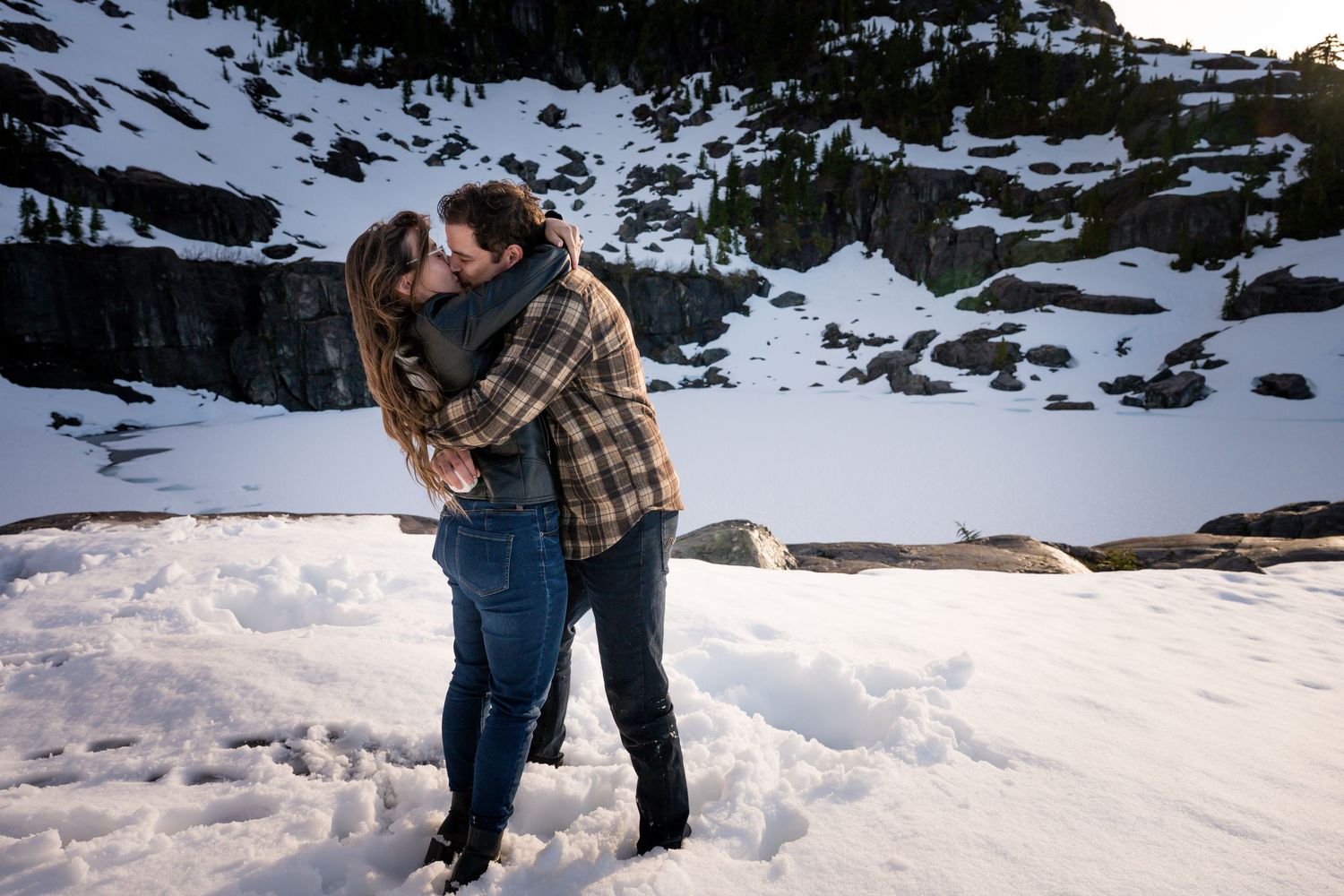 Couple kissing in snowy mountain landscape wearing casual winter clothing.