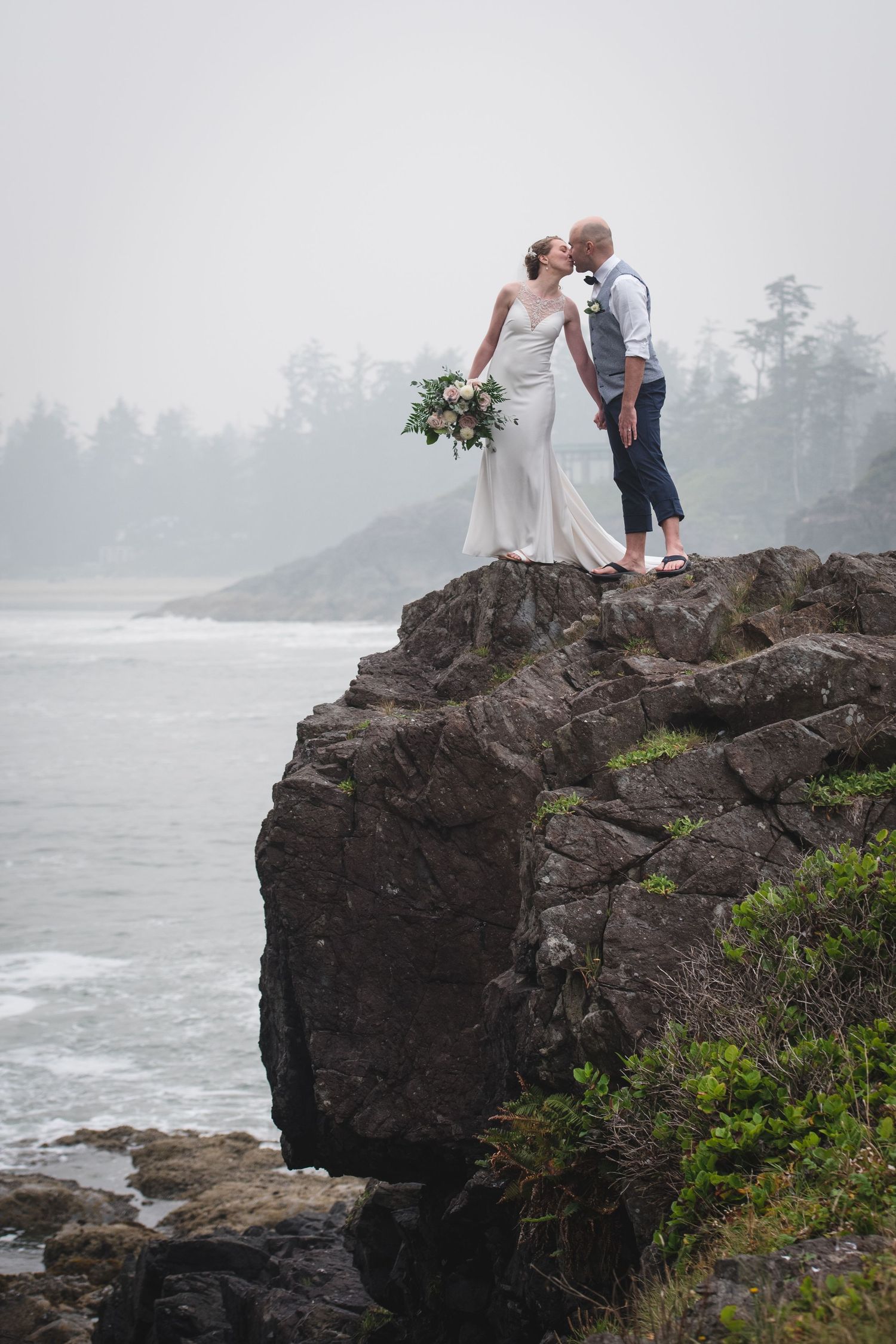 A wedding couple stands dramatically on a rocky cliff overlooking a misty Pacific Northwest coastline.