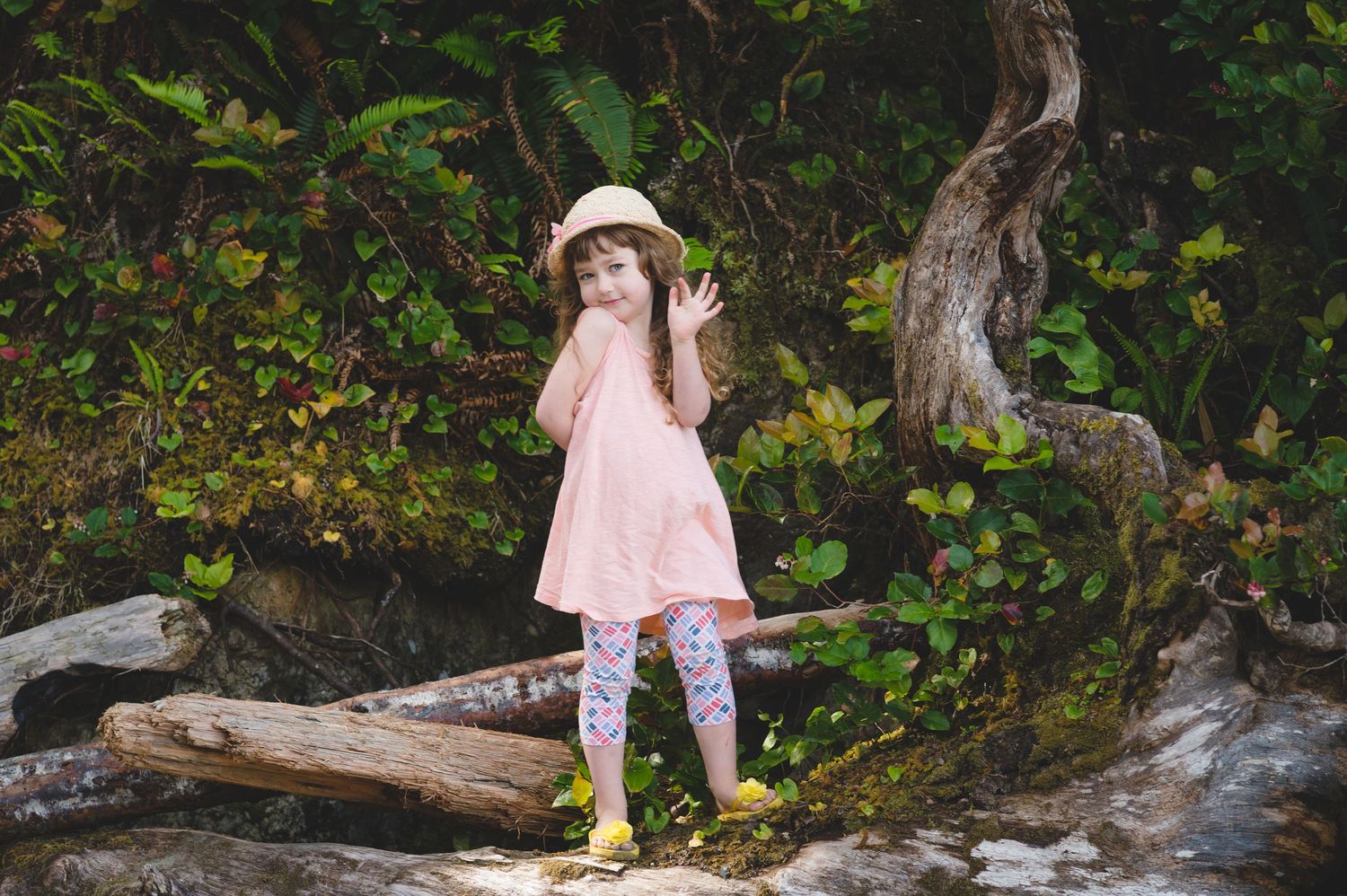 Young child in a pink dress explores a fallen log in a lush forest setting.
