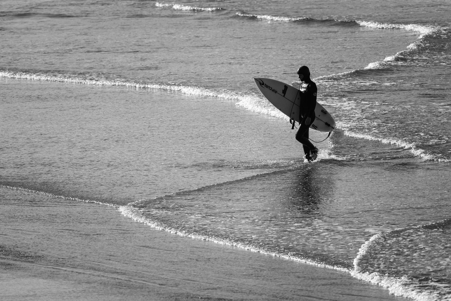 A silhouetted surfer walks along the shoreline with their surfboard in black and white photography.