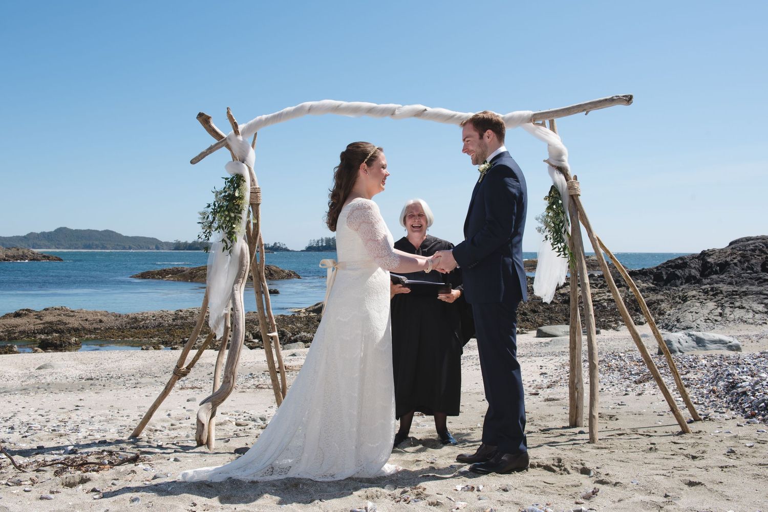 A beach wedding ceremony setup with driftwood arch and white chairs against a scenic ocean backdrop.