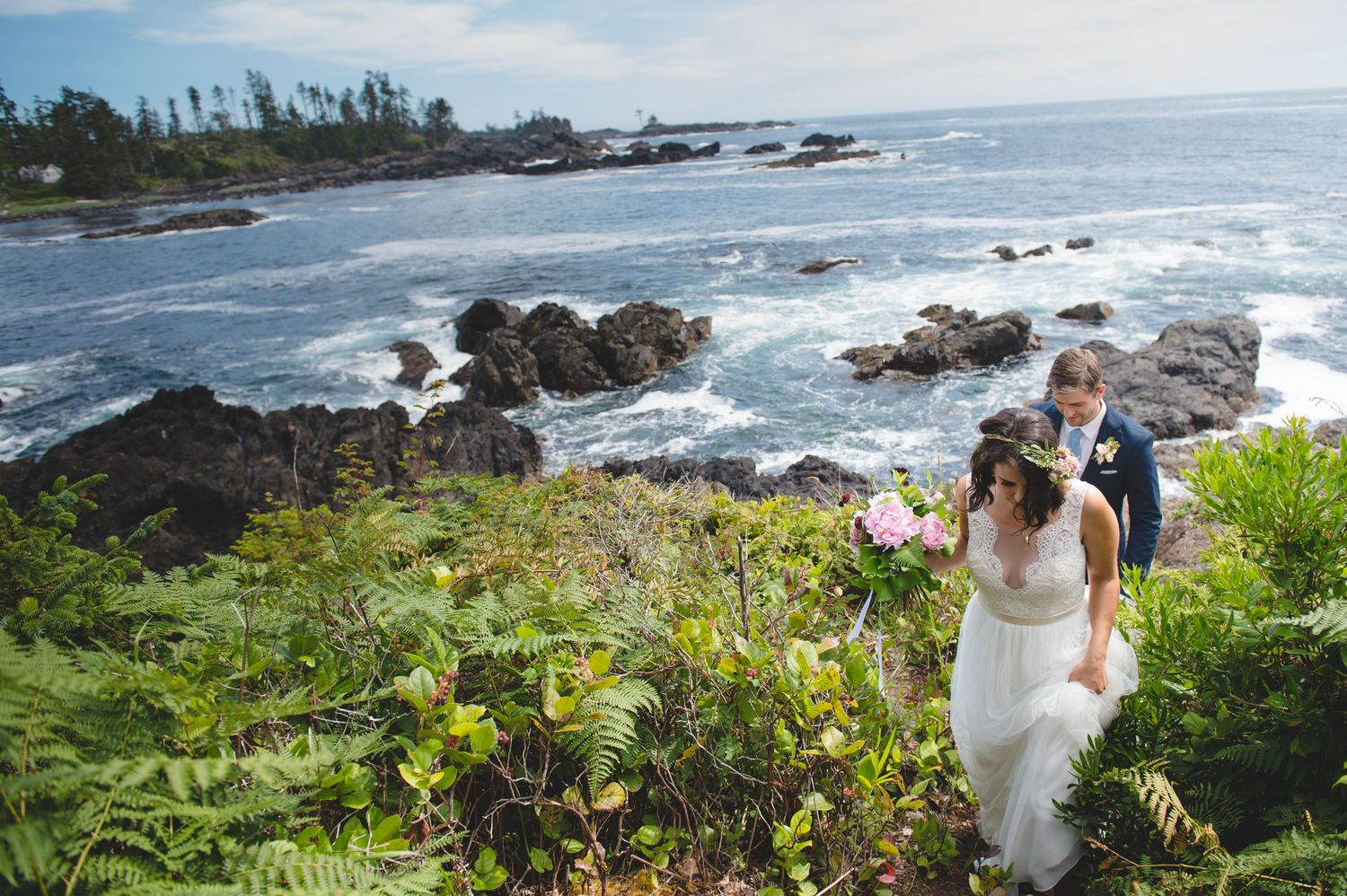 A couple walks along rugged ocean coastline with waves crashing against rocks below.