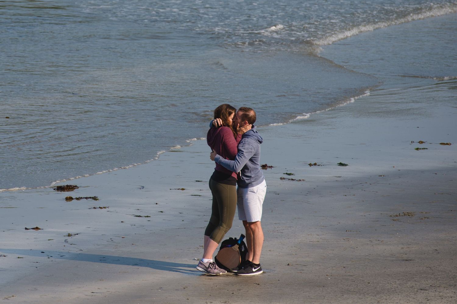 A person crouches down to observe something on a sandy beach with gentle waves in the background.
