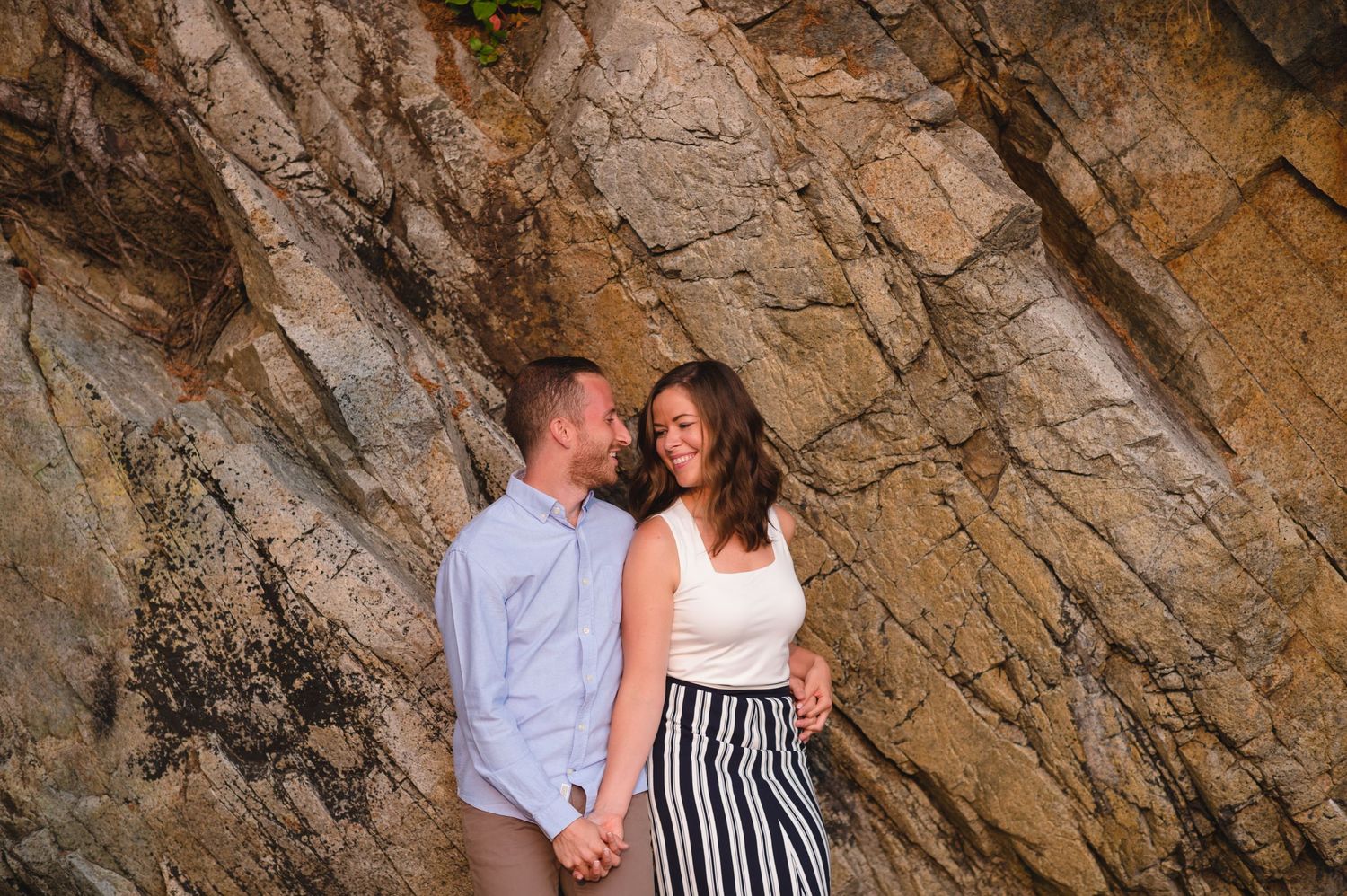 Couple stands close together against dramatic rocky cliff face during outdoor photo session.