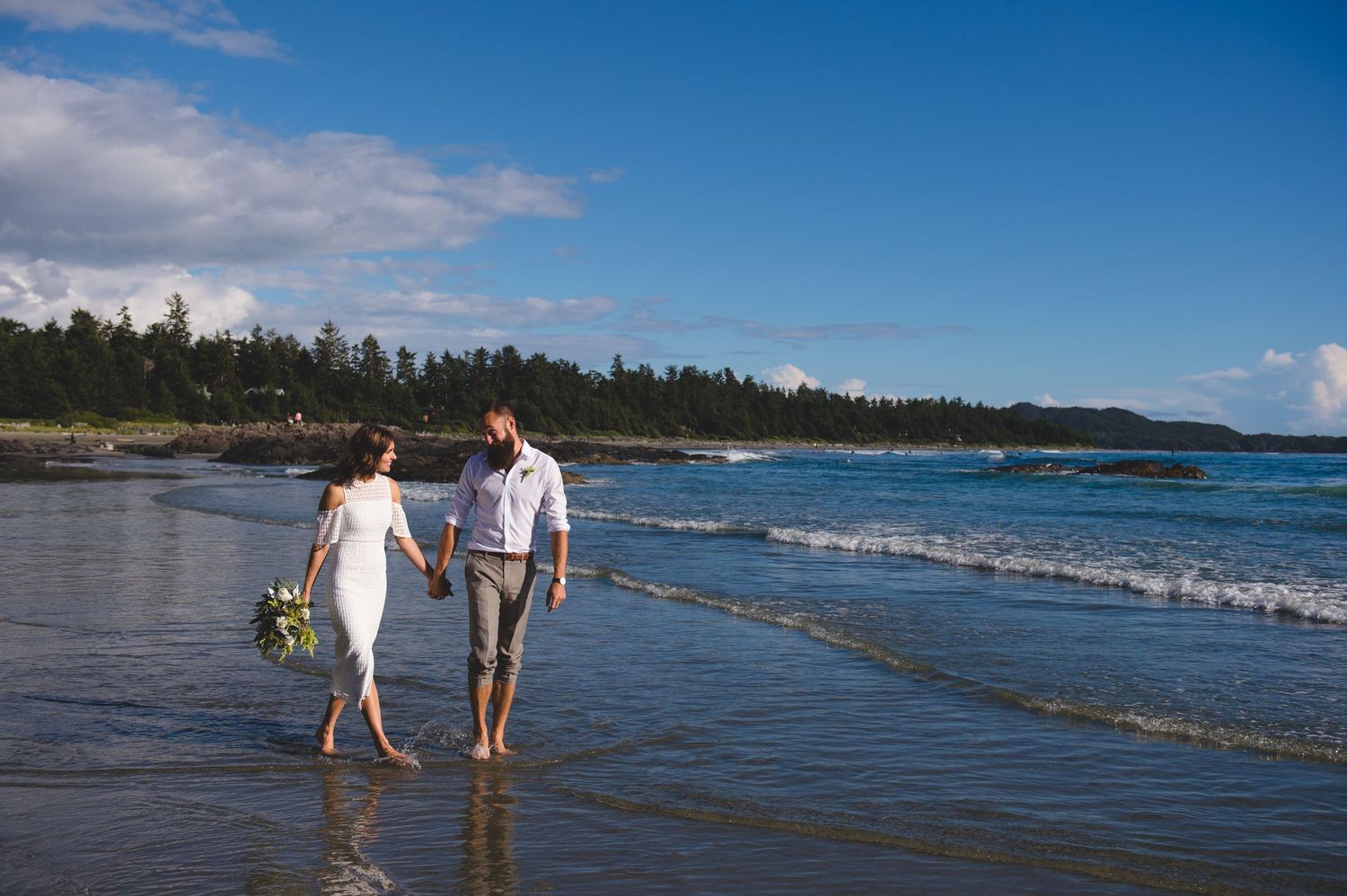Couple walking along scenic Pacific Northwest beach with waves and forested coastline on sunny day.
