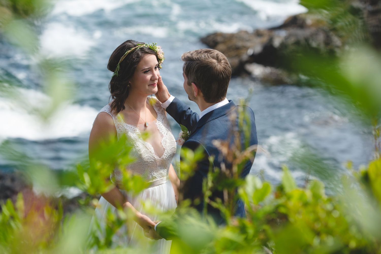 A couple shares an intimate moment by a rushing stream surrounded by green foliage on their wedding day.