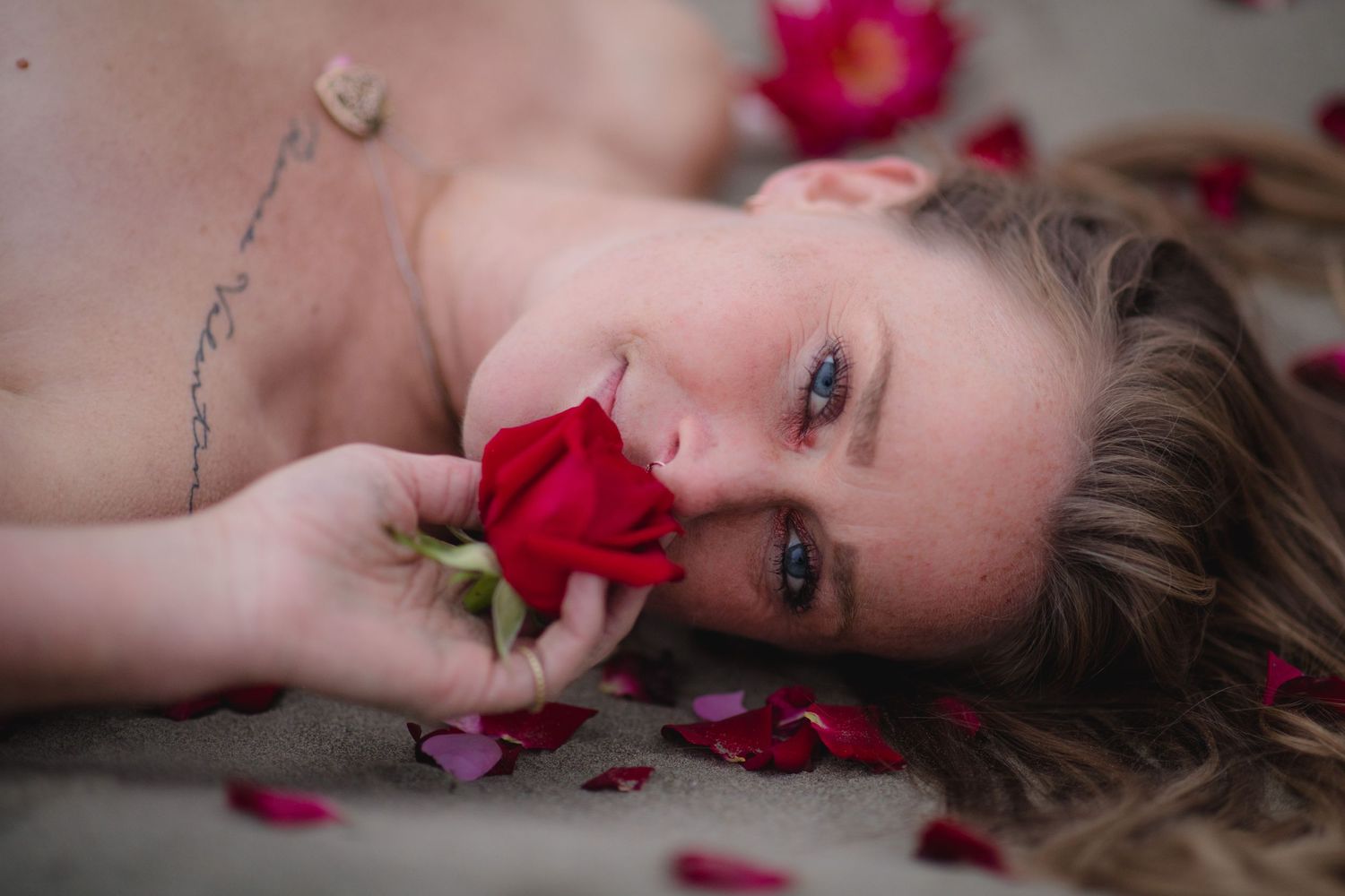 Red rose petals scattered around someone lying on white fabric in an artistic portrait setting.