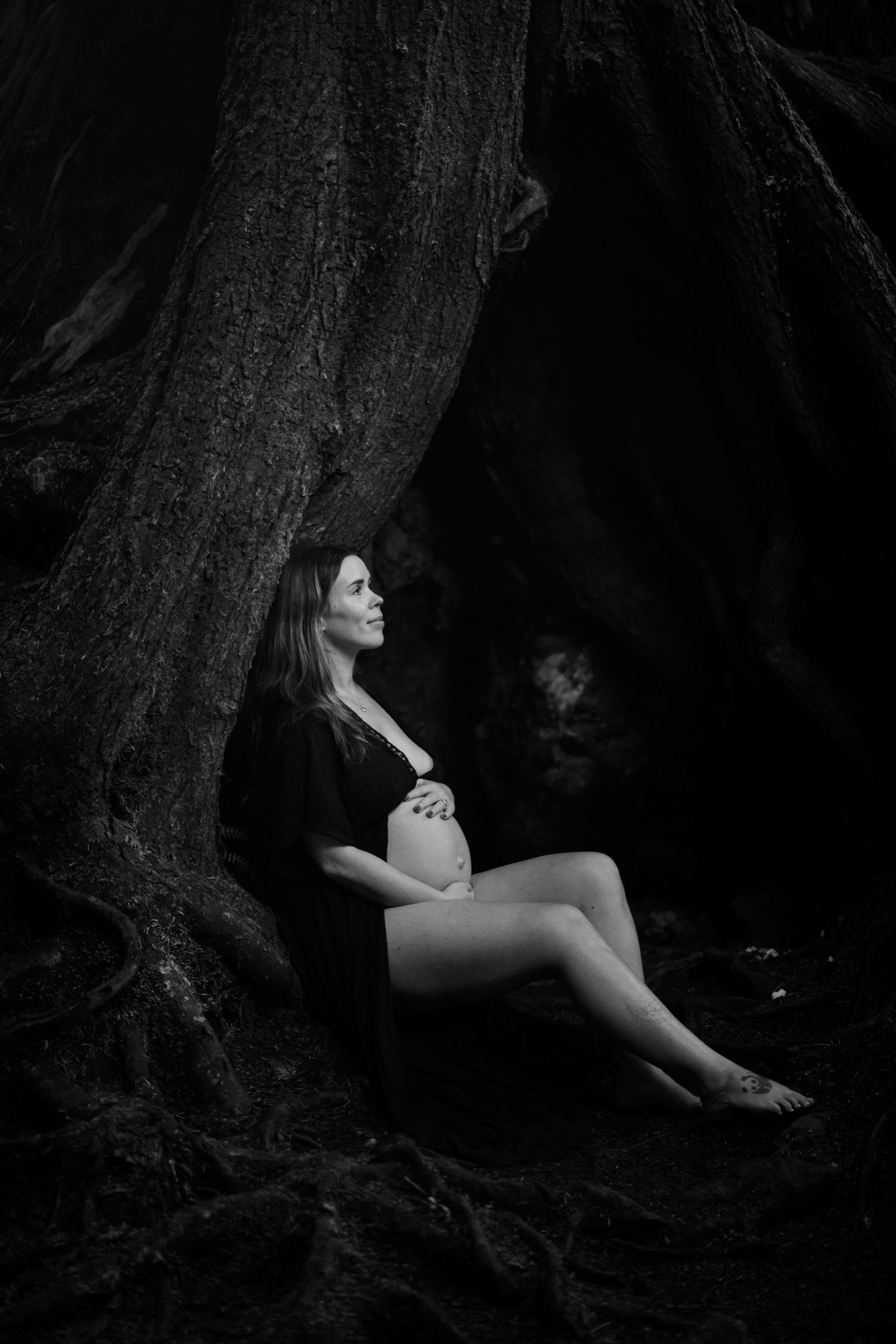 Black and white portraits of a person sitting against a large tree trunk in a moody forest setting.