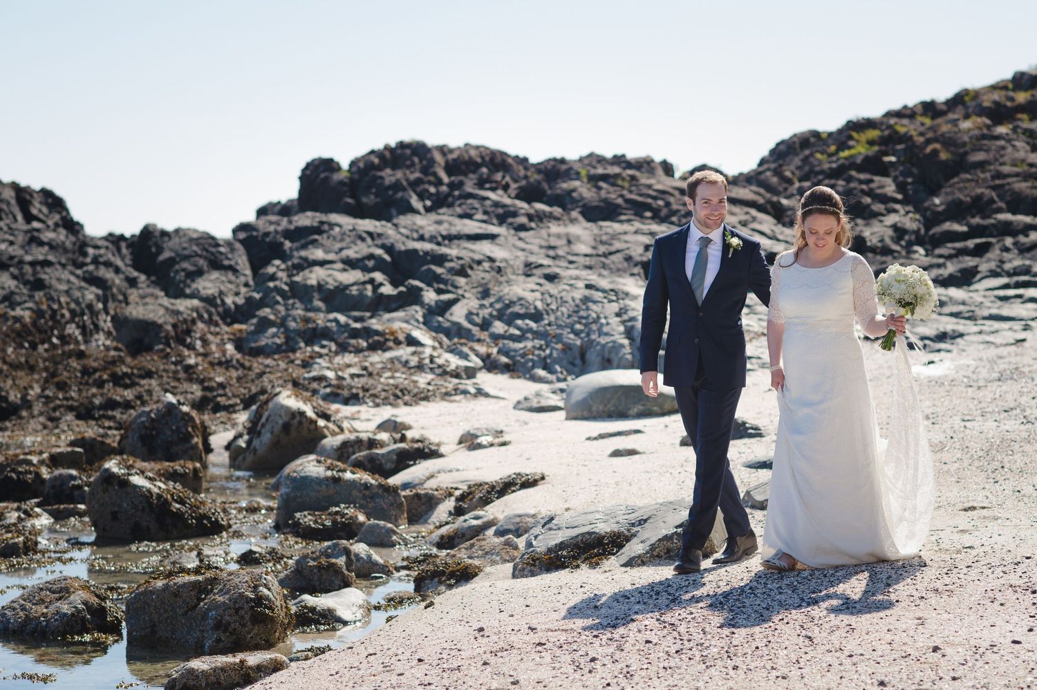A couple walks together on rocky coastal terrain wearing wedding attire.