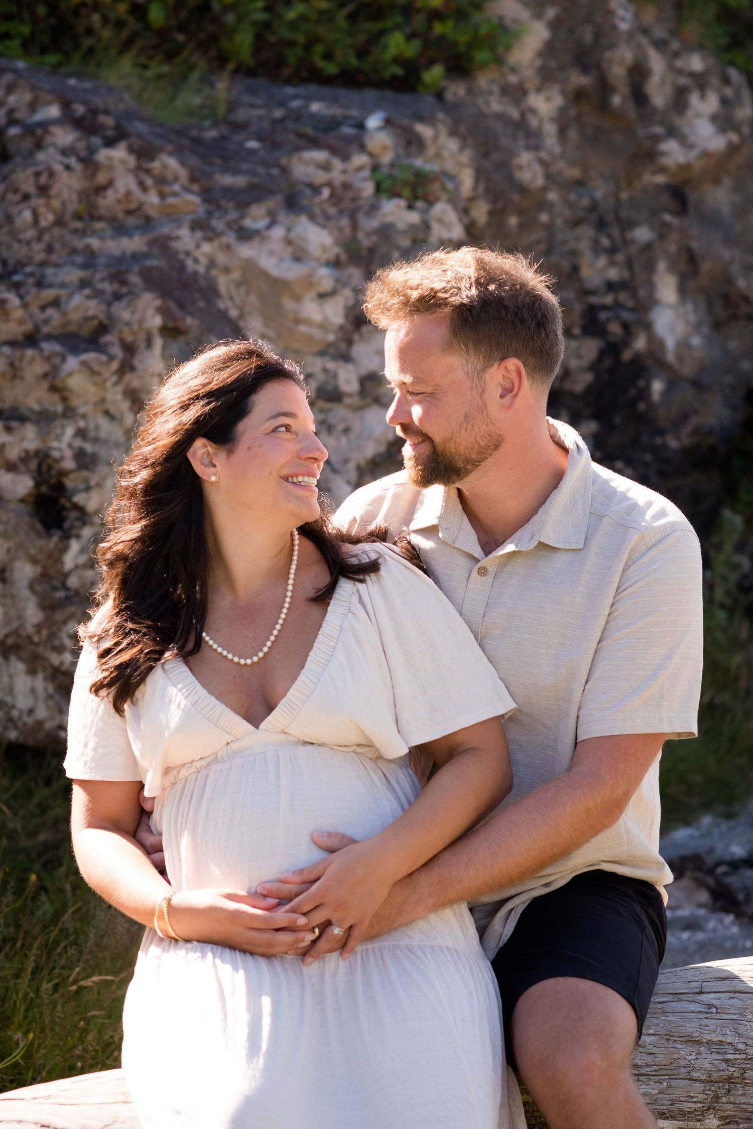 A couple in white tops shares loving glances while sitting on driftwood near rocky cliffs.