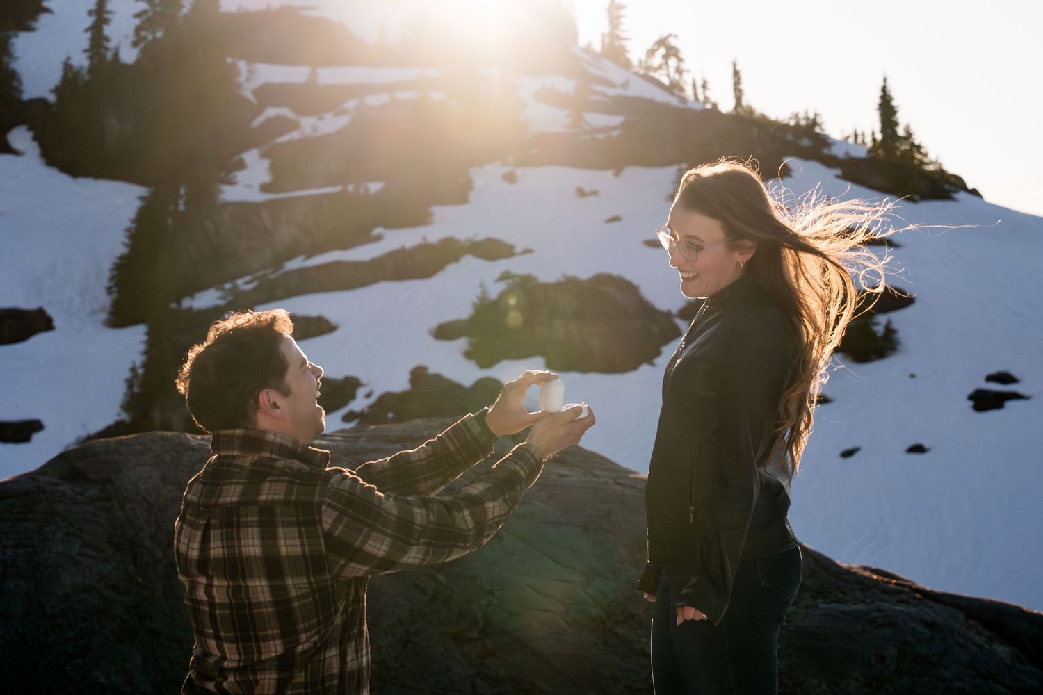 Man proposing on one knee to his girlfriend on a snowy mountain at golden hour with ring box.