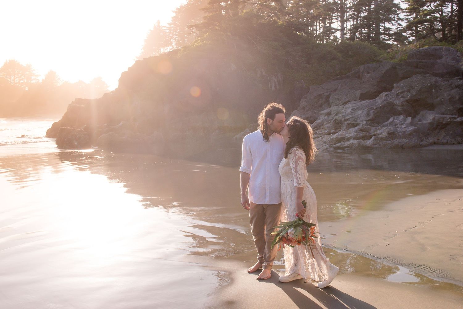 Couple sharing a romantic moment on a sunlit beach at golden hour with rocky cliffs and ocean in the background.