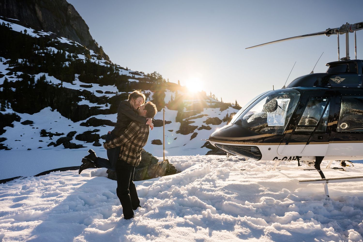 Couple embracing passionately in deep snow beside a helicopter during a mountain engagement session in Tofino.