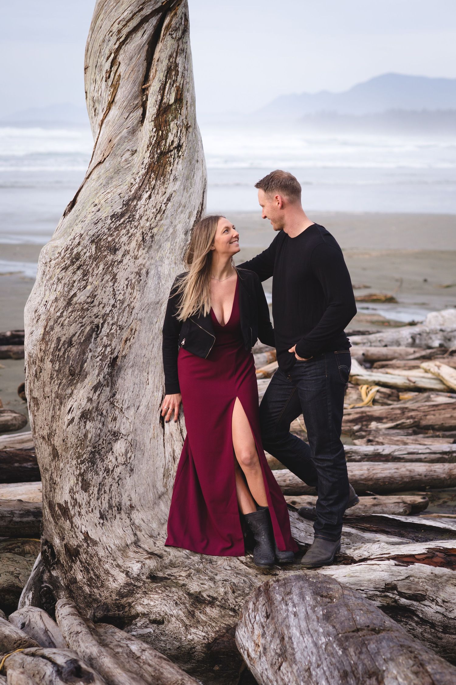 Couple in burgundy dress and black attire share intimate moments on driftwood at misty Pacific Northwest beach.