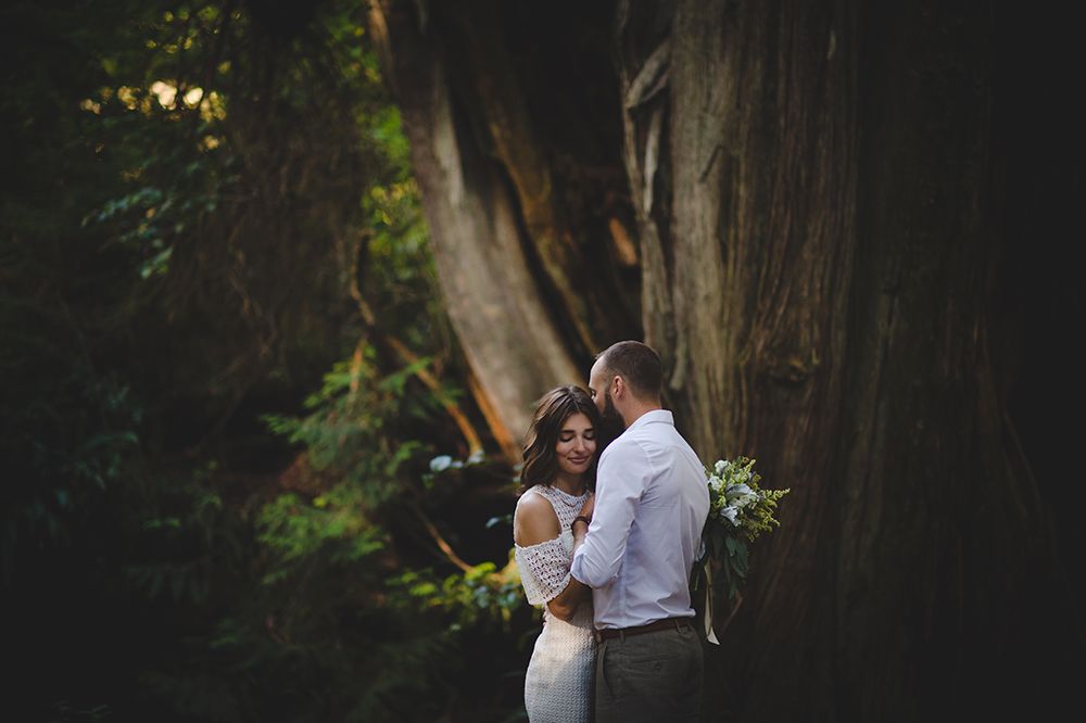 A couple embraces during their engagement photoshoot in a sunlit forest setting with dramatic lighting.
