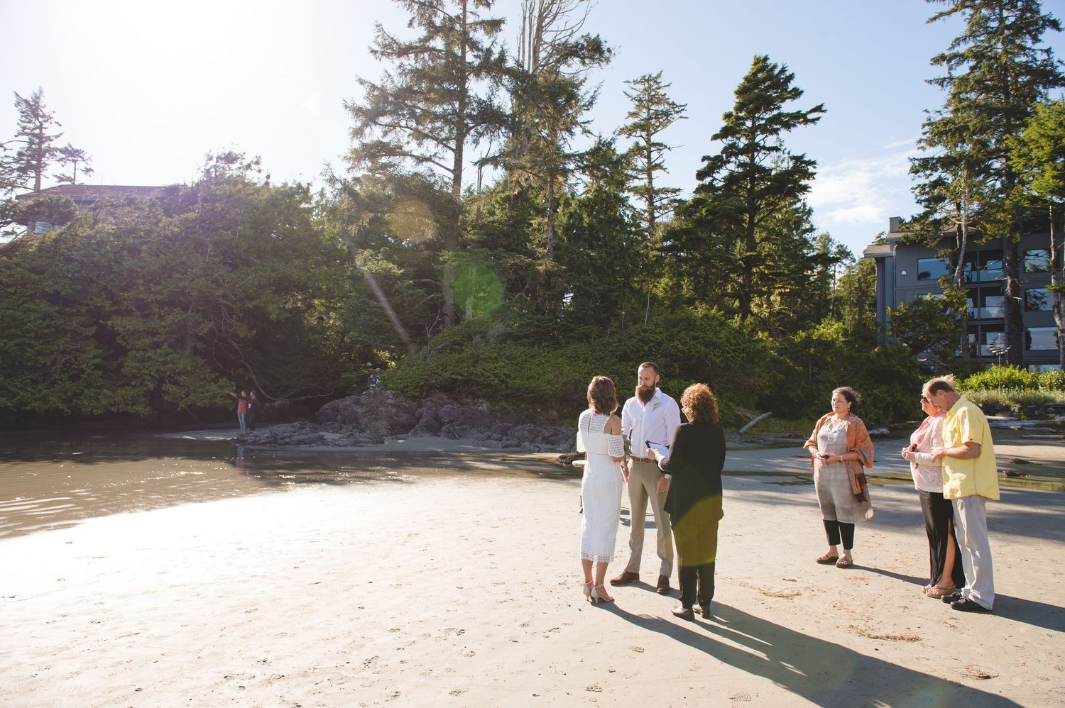 Wedding ceremony taking place on sandy beach with ocean and trees in background.