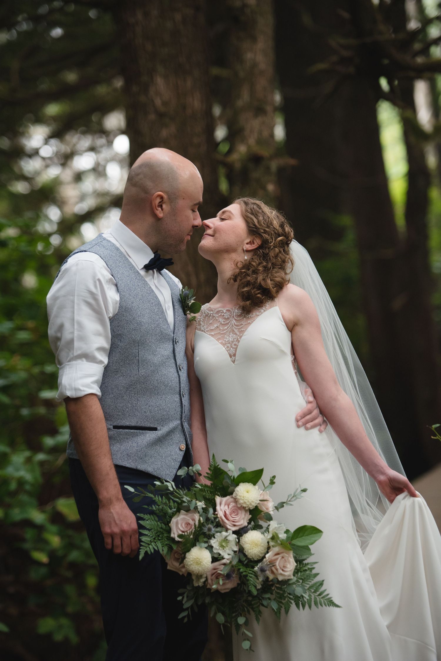 A romantic wedding couple shares tender moments in a forest setting while holding a delicate bouquet of roses and greenery.