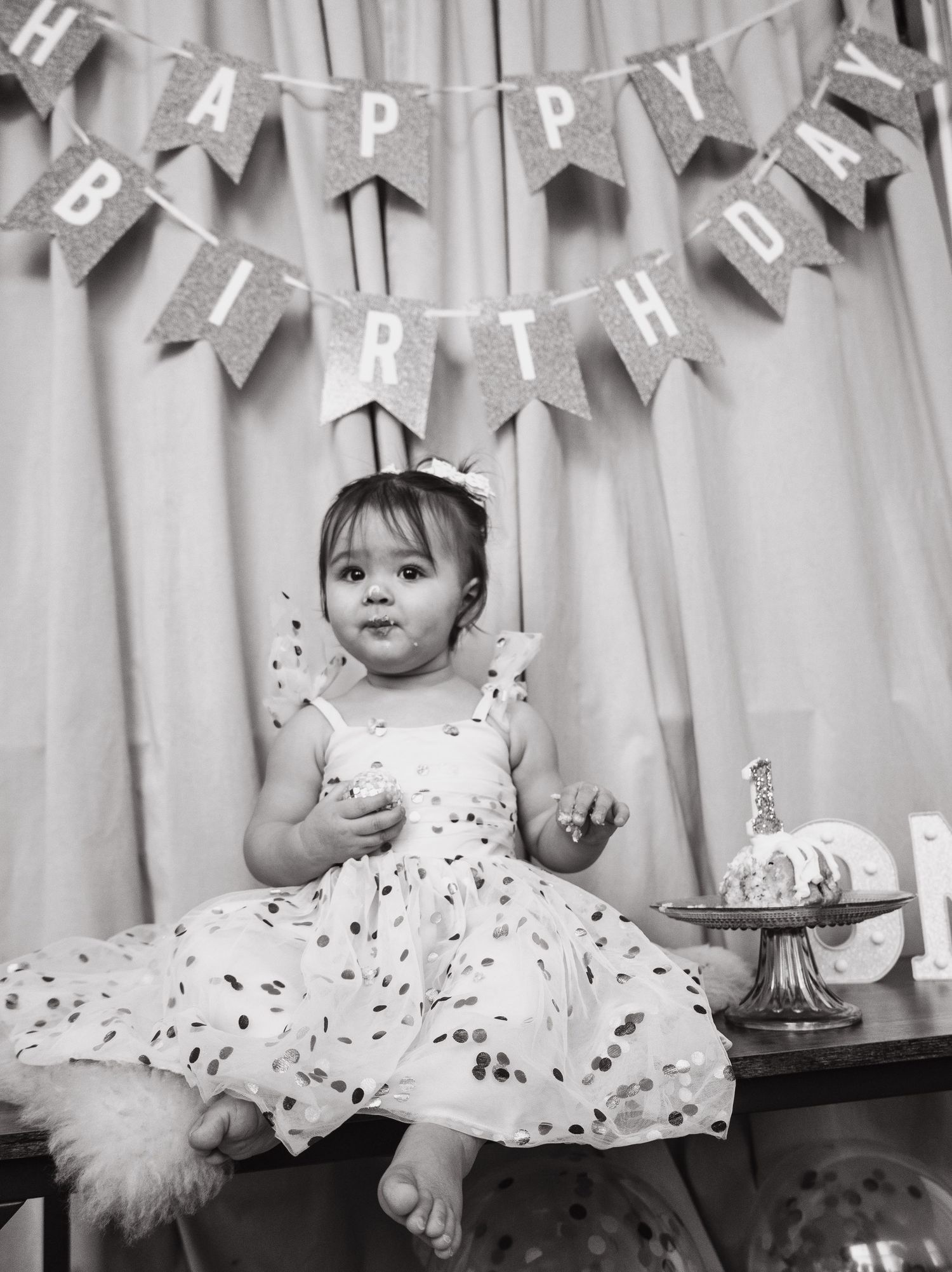 Baby in white dress sits under Happy Birthday banner with cake and decorations.
