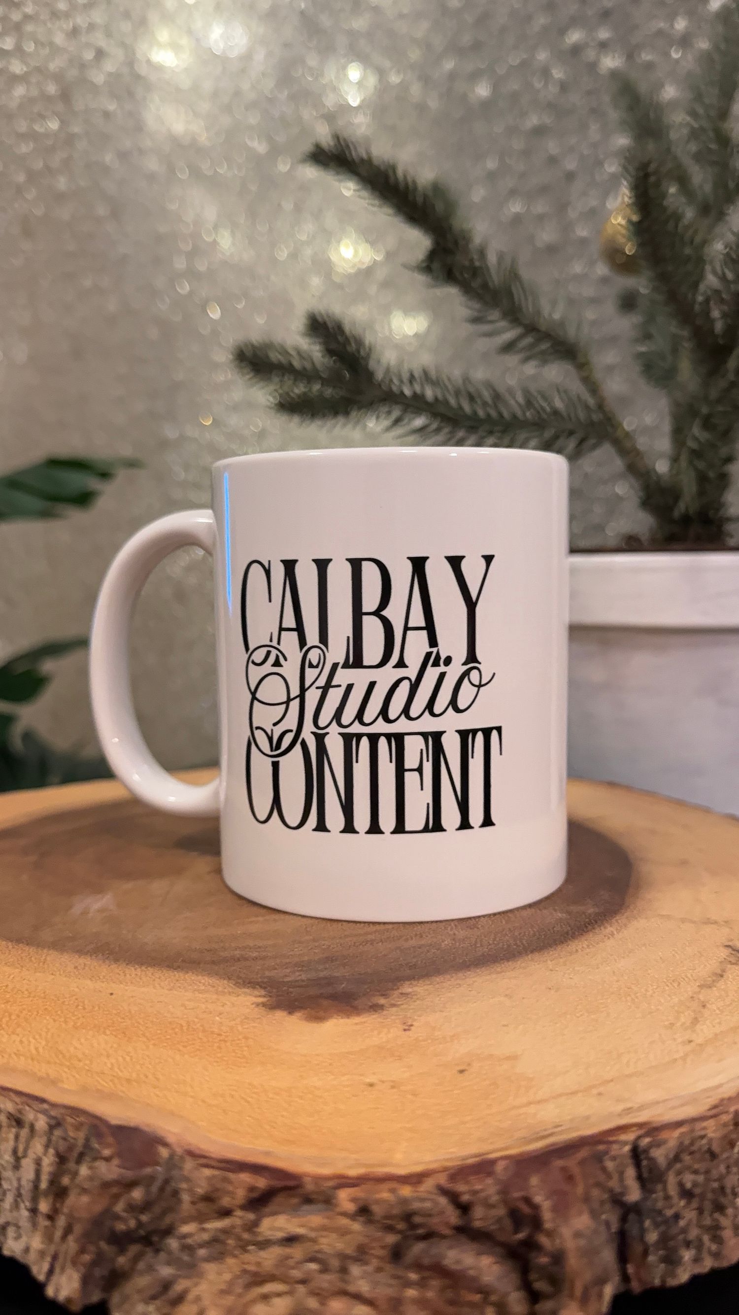 White ceramic coffee mug with modern black typography displayed on a wooden slice with evergreen branches in background.