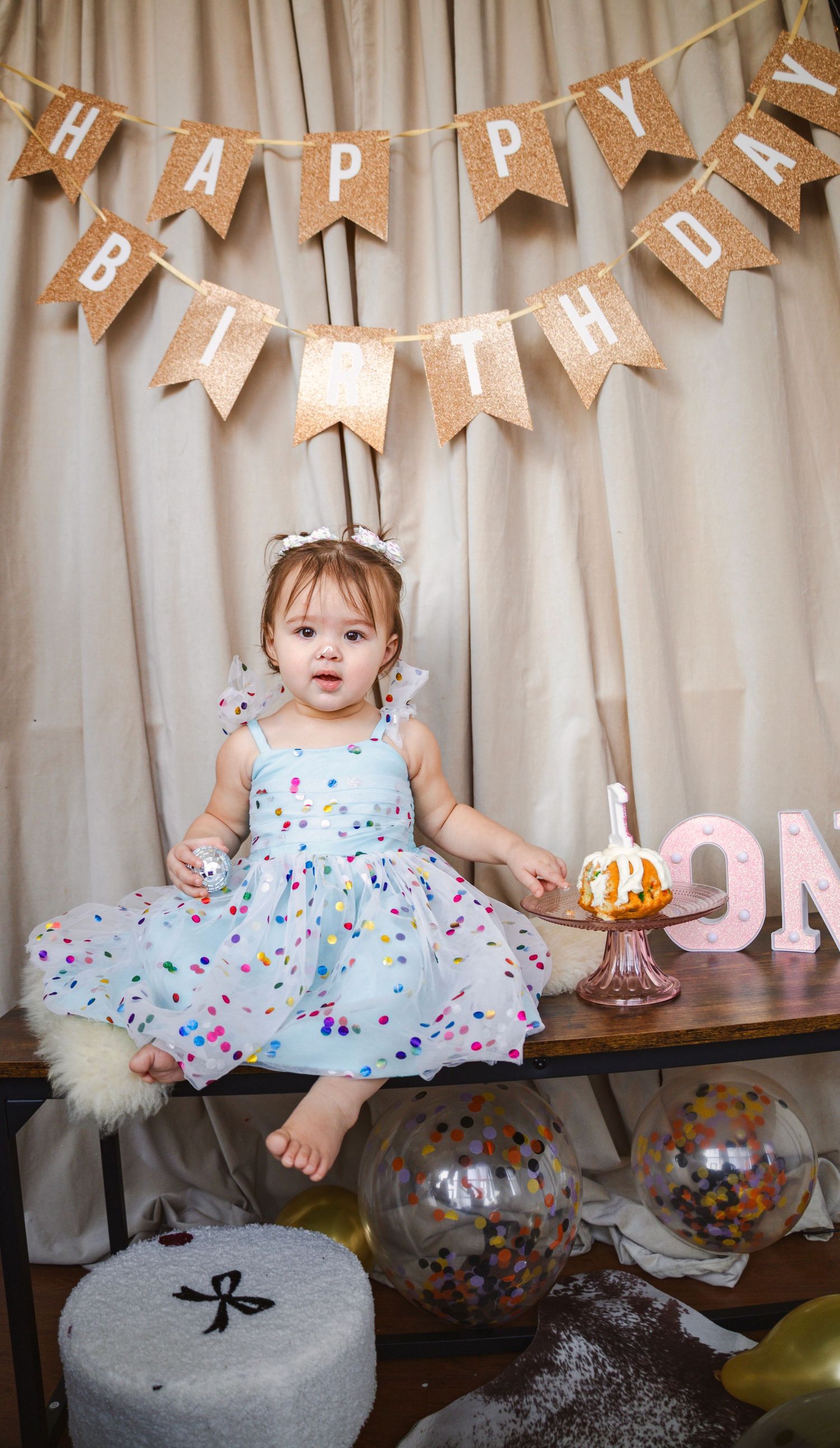 Baby in polka dot dress celebrates first birthday with cake and rustic banner decorations.